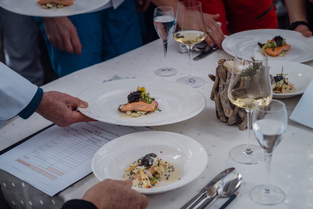 Essen wird serviert. Zwei Teller und Gläser Wein stehen am Tisch.